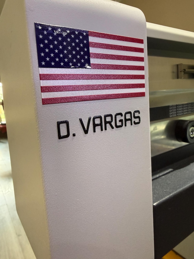 May include: A white machine surface featuring a rectangular sticker of the United States flag positioned above the black text D. VARGAS. The machine has a smooth, matte finish and a curved edge, with metallic components visible in the background.