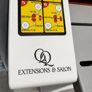 May include: A white machine panel featuring a black logo with the text "CQ EXTENSIONS & SALON" and a yellow instructional diagram labeled with numbers 5 and 6, illustrating the path of film and card paper through top and down rods and rolls.