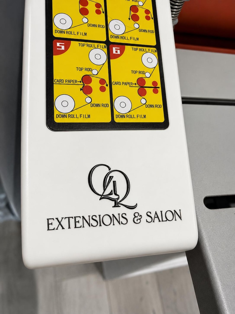 May include: A white machine panel featuring a black logo with the text "CQ EXTENSIONS & SALON" and a yellow instructional diagram labeled with numbers 5 and 6, illustrating the path of film and card paper through top and down rods and rolls.