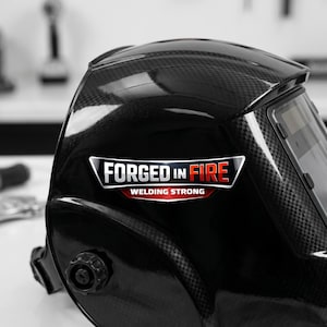 May include: A black welding helmet with a carbon fiber texture and a glossy finish. The side features a graphic decal with the text FORGED IN FIRE and WELDING STRONG in white, red, and silver lettering. The helmet includes an adjustable side knob.