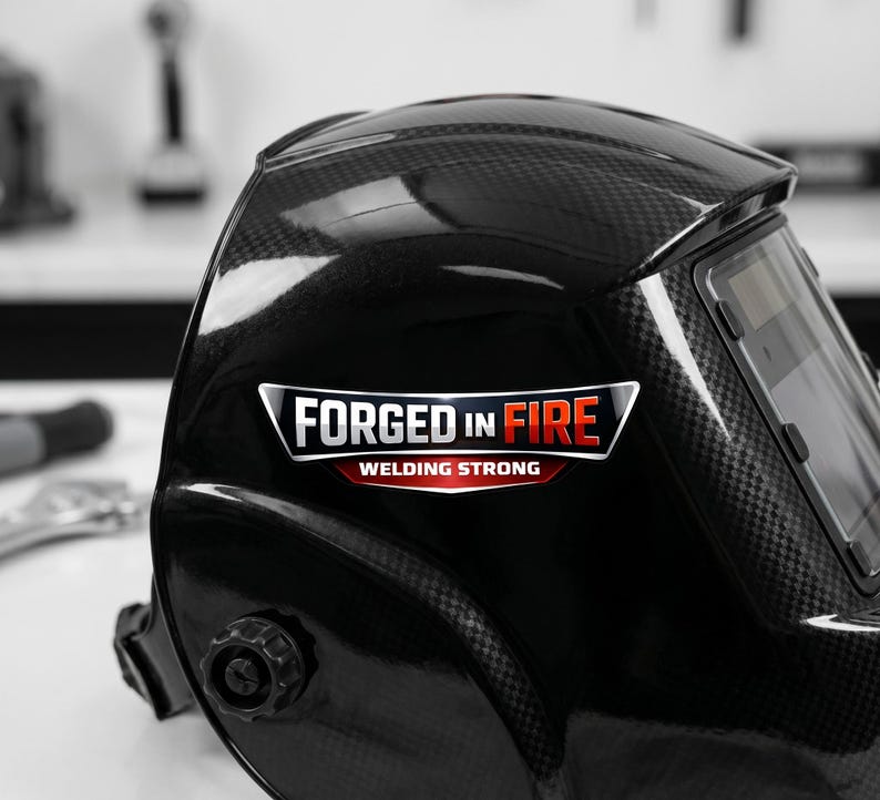 May include: A black welding helmet with a carbon fiber texture and a glossy finish. The side features a graphic decal with the text FORGED IN FIRE and WELDING STRONG in white, red, and silver lettering. The helmet includes an adjustable side knob.