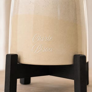 May include: A cylindrical ceramic vessel with a two-tone cream and beige speckled finish, featuring the engraved text "Cherie Bisou" on the front. The vessel is supported by a black wooden stand with four legs arranged in a cross pattern.