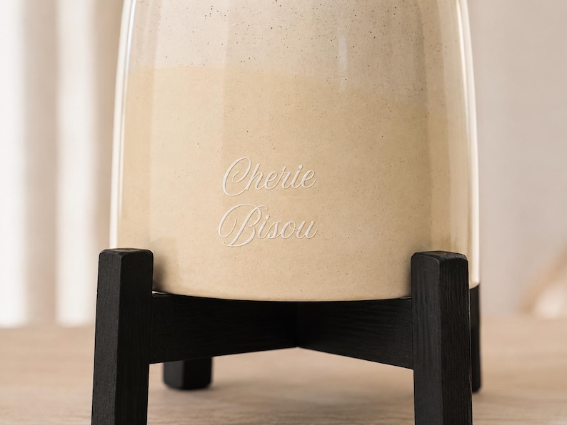 May include: A cylindrical ceramic vessel with a two-tone cream and beige speckled finish, featuring the engraved text "Cherie Bisou" on the front. The vessel is supported by a black wooden stand with four legs arranged in a cross pattern.