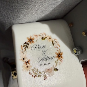 May include: A white surface featuring a printed floral wreath design in shades of beige, brown, and green. Inside the wreath, the text reads Rosa y Arturo with the date 09.26.26 printed below in a black script and sans-serif font combination.