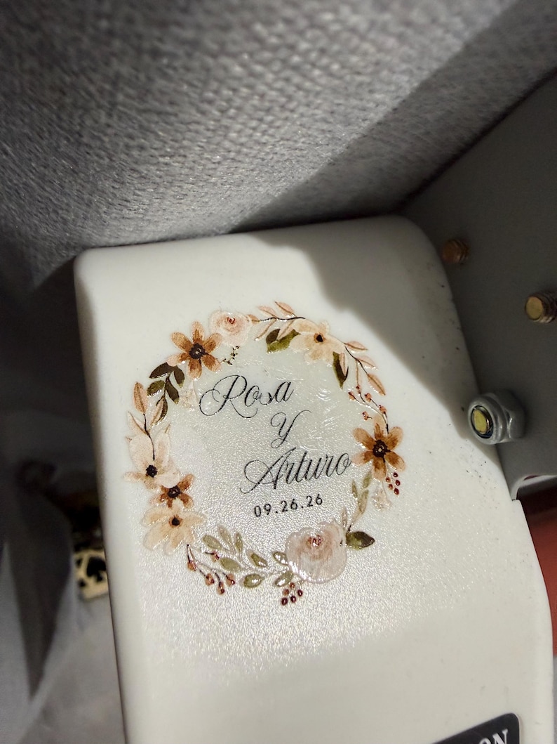 May include: A white surface featuring a printed floral wreath design in shades of beige, brown, and green. Inside the wreath, the text reads Rosa y Arturo with the date 09.26.26 printed below in a black script and sans-serif font combination.
