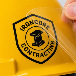 May include: A bright yellow hard hat featuring a black shield-shaped decal with the text IRONCORE CONTRACTING and an I-beam icon. A clear adhesive film is being peeled back from the surface of the sticker, revealing the glossy finish underneath.