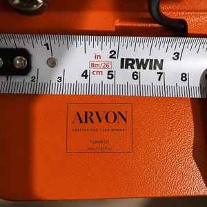 May include: An orange surface featuring a black rectangular transfer sticker with the text ARVON, CRAFTED FOR YOUR MOMENT, Cuticle Oil, and 10ml / 0.33 fl oz. An Irwin measuring tape shows the sticker width is approximately 1.5 inches wide.