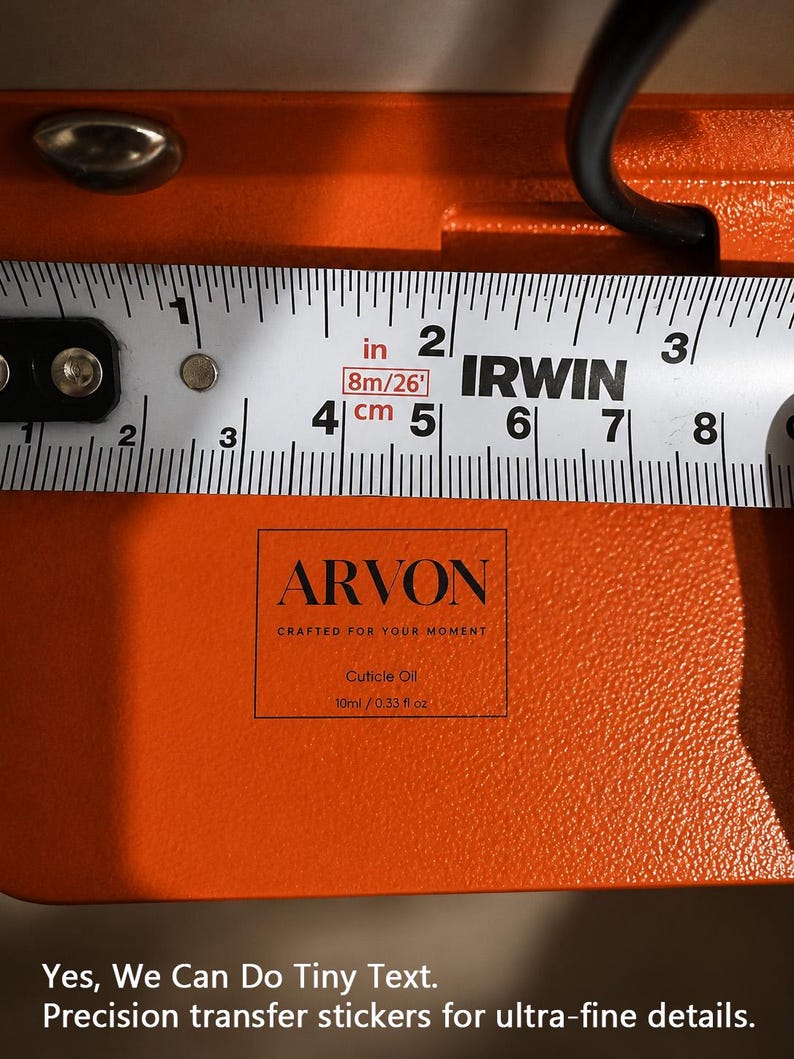 May include: An orange surface featuring a black rectangular transfer sticker with the text ARVON, CRAFTED FOR YOUR MOMENT, Cuticle Oil, and 10ml / 0.33 fl oz. An Irwin measuring tape shows the sticker width is approximately 1.5 inches wide.