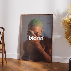 Puede incluir: Impresión artística enmarcada que presenta a una persona con cabello verde cubriéndose la cara. La palabra "blond" está impresa en blanco. La obra de arte se exhibe en una habitación con piso de madera y una pared de color neutro.