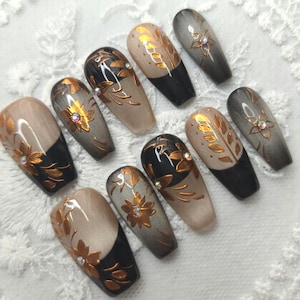 May include: A set of ten artificial nails with a floral design. The nails feature a combination of black, beige, and gray colors, with gold floral accents and small rhinestones. The nails are arranged on a white lace surface.