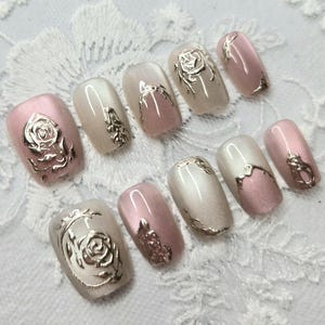 May include: A set of ten artificial nails in various shapes and sizes. The nails are painted in shades of pink and white, with silver rose and floral designs. The nails are arranged on a white lace surface.