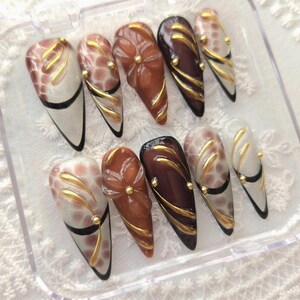 May include: A set of ten almond-shaped false nails in a clear plastic case. The nails feature various designs, including gold swirls, floral accents, and a mix of brown, white, and black colors. Each nail has a glossy finish.