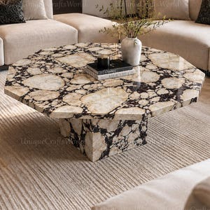 Op de afbeelding: Een achthoekige salontafel met een gepolijst oppervlak met een crème, beige en donkerbruin breccie-steenpatroon. De tafel wordt ondersteund door een centrale vierkante voet van hetzelfde steenmateriaal, wat zorgt voor een samenhangende uitstraling.
