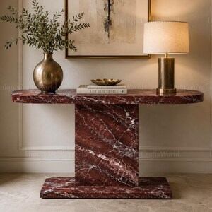 May include: A dark red marble console table with a smooth oval top and a rectangular pedestal base. The table features prominent white veining throughout the stone surface. The piece is displayed with a brass vase, a small bowl, and a lamp.