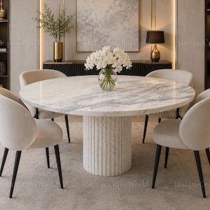 Pode incluir: Uma mesa de jantar redonda com tampo de mármore branco e base cilíndrica canelada, rodeada por quatro cadeiras estofadas em tecido bege claro com pernas pretas. Um vaso de vidro com flores brancas está colocado no centro da mesa.