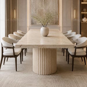 Op de afbeelding: Een rechthoekige eettafel met een dik, lichtbeige stenen blad en een cilindrische, gecanneleerde voet. Acht stoelen met donkerhouten frames en wit gestoffeerde zittingen staan rond de tafel. Een witte keramische vaas met takken staat in het midden.