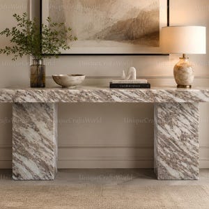 May include: A rectangular console table featuring a white and brown marble pattern with two thick, block-style legs. The table surface holds a glass vase with green foliage, a marble bowl, a white abstract sculpture, and a lamp with a beige shade.