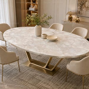 May include: An oval dining table with a white marble-patterned top and a gold-toned metal base, surrounded by four beige upholstered chairs with gold-toned metal legs. A white ribbed vase and two small decorative bowls sit on the table surface.