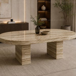 Peut inclure: Une table à manger ovale fabriquée en pierre de travertin beige polie avec une surface lisse et brillante. La table présente un plateau épais et solide soutenu par deux pieds rectangulaires, mettant en valeur le veinage naturel de la pierre.