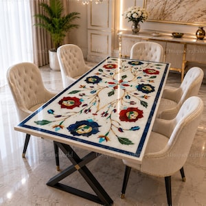 Op de afbeelding: Een rechthoekige eettafel met een wit marmeren blad met een ingelegd bloemmotief van rode, blauwe en groene steen. De tafel heeft een zwart metalen X-vormig onderstel en is omringd door zes beige gestoffeerde stoelen met gecapitonneerde rug.