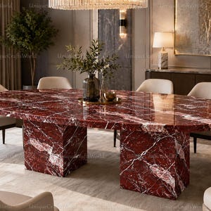 Op de afbeelding: Een ovale eettafel met een gepolijst dieprood marmeren blad met witte aders, ondersteund door twee bijpassende rechthoekige marmeren voetstukken, omringd door zes beige gestoffeerde stoelen met donkere houten poten.