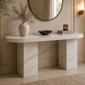May include: An oval console table crafted from white marble with grey veining, featuring two thick rectangular pillar legs. The tabletop holds a beige ceramic vase with greenery, a shallow black bowl, and a stack of two books with a candle.