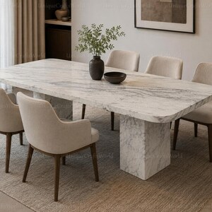 Peut inclure: Une table à manger rectangulaire avec un plateau en marbre blanc et un pied central en marbre assorti, accompagnée de six chaises rembourrées en tissu beige clair avec des pieds en bois foncé. Un vase sombre et un bol sont posés dessus.