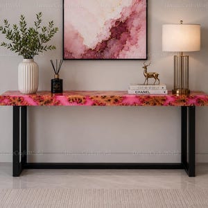 Può includere: Un tavolo console rettangolare caratterizzato da un piano in resina rosa e oro con venature del legno naturali, sostenuto da una robusta struttura in metallo nero. La superficie del tavolo presenta una finitura lucida con dettagli colorati.