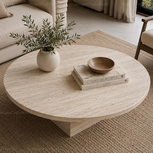 May include: A round beige travertine coffee table with a textured stone surface and a pedestal base. On the table sits a white ceramic vase with green foliage, a small wooden bowl, and two stacked books titled Live Beautiful at 100.