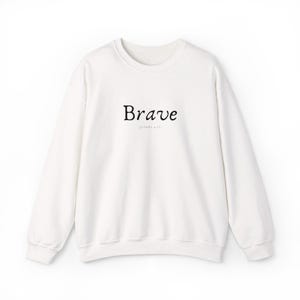 Op de afbeelding: Een witte crew neck sweatshirt met het woord "Brave" in zwart schrift over de borst. De tekst "Jonah 1:13" is eronder gedrukt. De sweatshirt heeft lange mouwen en een relaxte pasvorm.