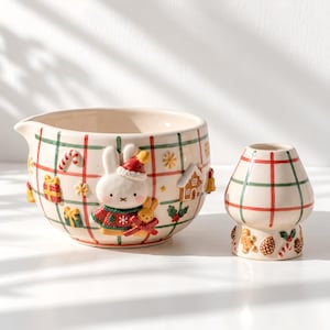 以下が含まれることがあります： クリーム色のベースに赤と緑のチェック柄が施された陶器製のミキシングボウルと小さなピッチャー。ボウルには、赤い帽子とセーターを着た白いウサギの立体的な装飾に加え、ジンジャーブレッドハウスやプレゼントのモチーフがあしらわれています。