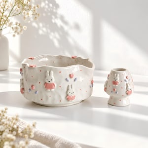 May include: A white ceramic bowl and a matching small vase, both featuring a textured surface with raised pink and white rabbit figures, small red polka dots, and pastel-coloured heart and leaf accents. The bowl has a wavy, uneven rim.