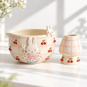 May include: A ceramic mixing bowl and a matching small cup, both featuring a cream-colored base with a pink grid pattern and red cherry motifs. The bowl displays a raised white rabbit figure wearing a patterned kimono and holding a fan.