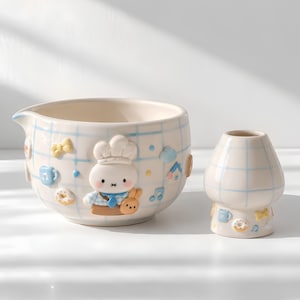 May include: A ceramic mixing bowl and a matching small vase, both featuring a light blue grid pattern on a cream background. The bowl displays a raised 3D character wearing a chef hat, surrounded by small embossed icons of bows, donuts, and musical notes.
