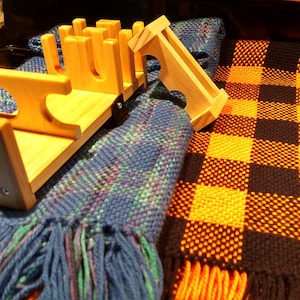 May include: A wooden knife block with a light brown finish. The block has slots for holding knives and a raised platform on the top. The block is sitting on a blue and green plaid blanket with orange and black checkered blanket in the background.