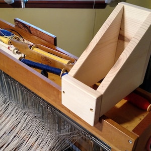 May include: A wooden loom with a wooden shelf attached to the side. The loom is set up with white yarn and there are spools of blue and yellow yarn in the loom's storage compartment. There are also spools of red yarn on the floor.