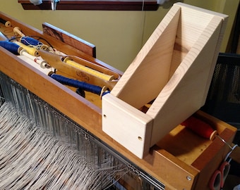 May include: A wooden loom with a wooden shelf attached to the side. The loom is set up with white yarn and there are spools of blue and yellow yarn in the loom's storage compartment. There are also spools of red yarn on the floor.