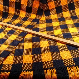 May include: A woven throw blanket with a black and orange checkered pattern. The blanket has fringe on the edges and is lying on a wooden surface.