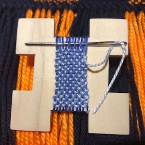 May include: A wooden loom with a blue and white woven tapestry. The loom is made of wood and has a white string attached to it. The tapestry is woven with blue and white yarn.