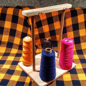 May include: A wooden yarn holder with four spools of yarn. The yarn is orange, black, blue, and pink. The yarn holder is on a checkered orange and black blanket.