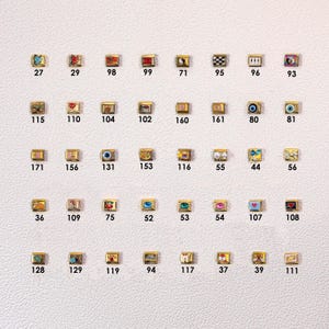 May include: A collection of small, gold-toned charms with various designs, including hearts, eyes, and geometric patterns. Each charm has a unique number, such as 27, 29, and 98, and is displayed on a light background.