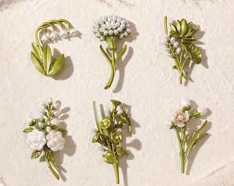 May include: A collection of six floral brooches featuring green metal stems and leaves accented with white faux pearls. The designs include lily of the valley, a carnation, a lotus, roses, morning glories, and a small blossom, all arranged in two rows.