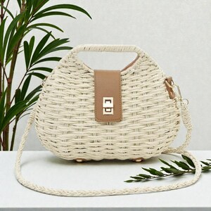 May include: Cream-colored woven handbag with a rounded shape and a top handle. The bag features a brown leather flap with a gold-colored clasp. A matching braided strap is attached for shoulder or crossbody wear. The bag has small gold feet.