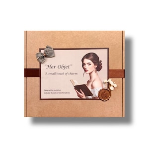 May include: A square cardboard box featuring a label with the text Her Objet, A small touch of charm, Designed by Journal up, Includes 18 packs of assorted pieces. The label shows an illustration of a person reading a book, a gingham bow, and a wax seal.