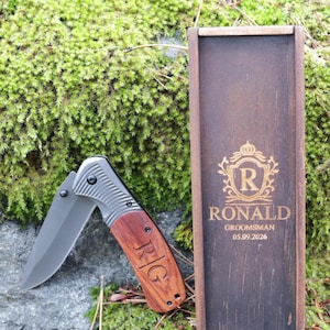 May include: A folding pocket knife with a wooden handle and a silver blade, next to a wooden box. The knife handle is engraved with the initials "R|G". The box is engraved with the name "RONALD", the word "GROOMSMAN", and the date "05.09.2026".