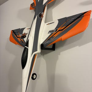 May include: A white, gray, and orange Horizon E-flite model airplane is mounted on a wall. The airplane has a sleek design with a black nose cone and a small wheel. The wings and tail feature orange accents.