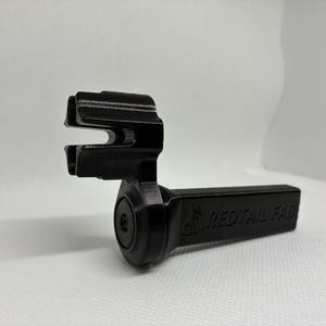 May include: A black, 3D-printed object with a unique shape, featuring a rectangular handle and a curved, slotted top. The text "REDTAIL FAB" is visible on the handle. The object is set against a white background.