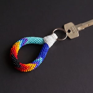 May include: A beaded keychain with a rainbow design, featuring black, red, orange, yellow, blue, and white beads. The keychain is attached to a silver key. The background is a dark gray surface.