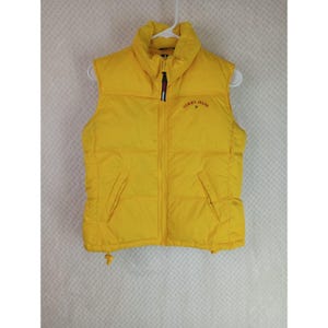 May include: A bright yellow puffer vest featuring a stand-up collar, a full-length front zipper closure, and two side pockets. The text Tommy Jeans is embroidered in small red and blue letters on the upper left chest area of the garment.