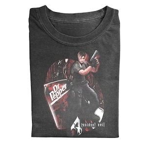 May include: Dark gray t-shirt featuring a graphic of a person holding a gun, with a Dr. Pepper can and the words "resident evil". The design is in shades of red, black, and white.